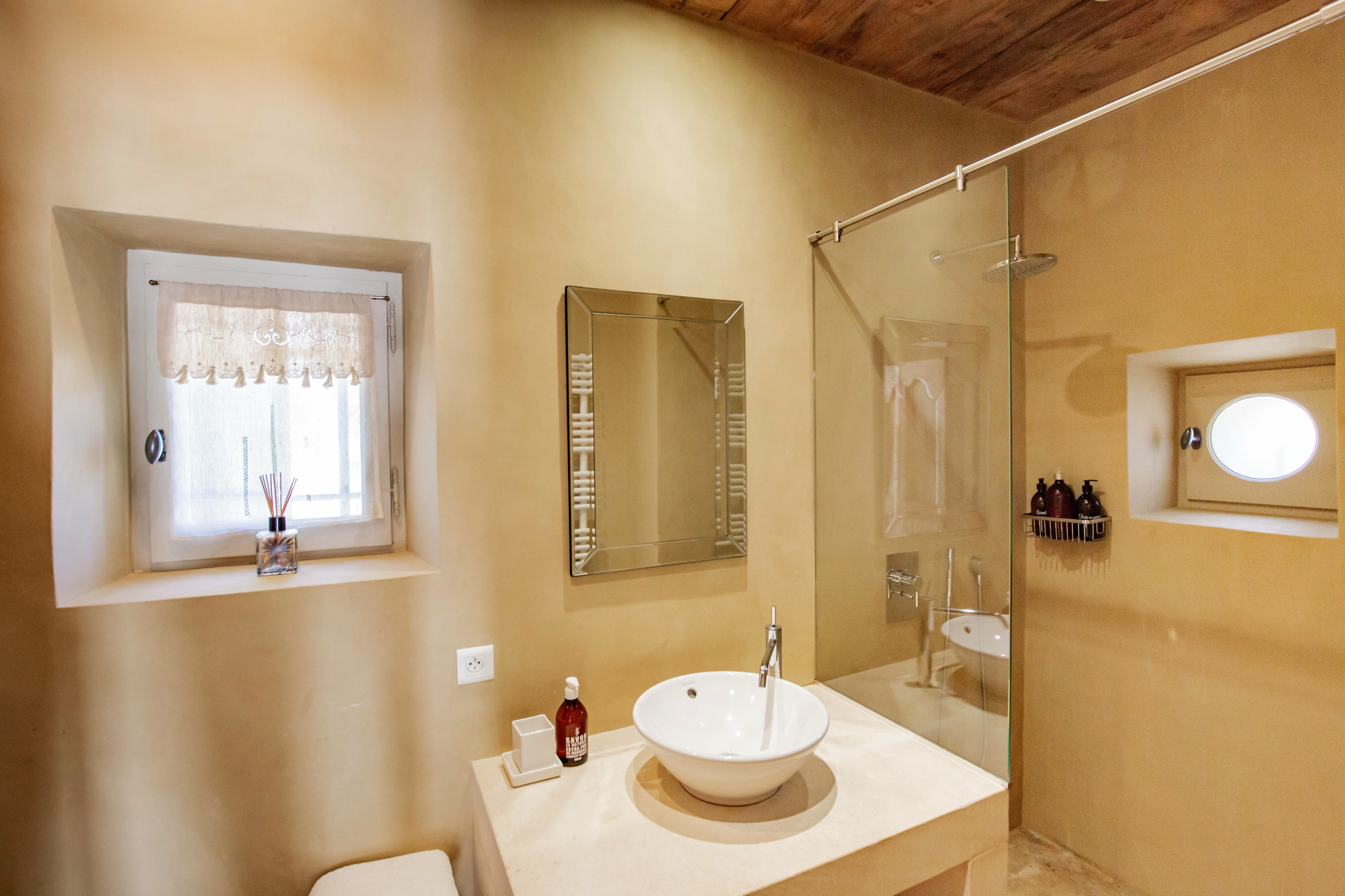 A warm-toned bathroom featuring a vessel sink, glass shower stall, and a window with white curtains. Neutral walls emphasize a bright and clean design.